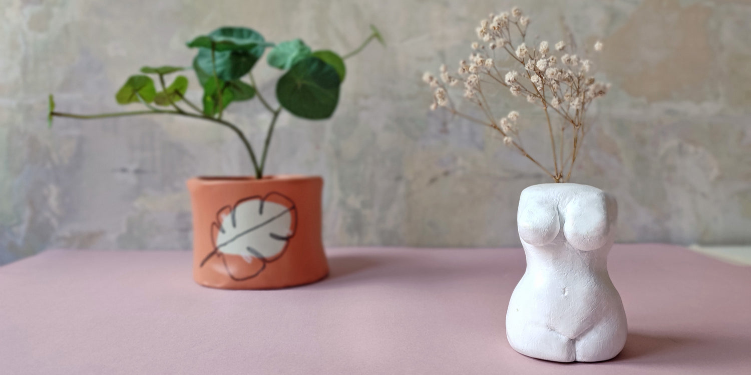 terrakotta színű agyag kaspó zöld virággal és egy női testet ábrázoló, levegőn száradó agyagból készült fehér szobor natúr száraz virággal, pasztell háttér előtt